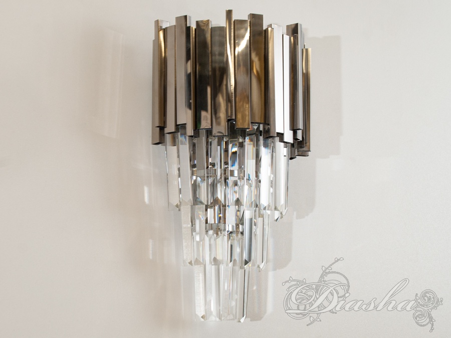 Сучасний кришталевий настінний світильник на дві лампи патрон Е14 представляє собою втілення елегантності та розкоші. Він складається з ретельно розташованих кришталів, що створюють вишукану композицію світла та відблисків, перетворюючи будь-яке приміщення в ексклюзивний простір.
Конструкція світильника включає в себе сміливе поєднання металу та скла, де матові та поліровані металеві елементи створюють контраст з прозорими кришталевими стрижнями, що падають каскадом. Металеві елементи мають сучасну обробку, яка гармонійно вписується в будь-який дизайнерський напрямок, від лофту до мінімалізму, додаючи кожному настінному світильнику унікальний характер.
Елегантний дизайн настінного світильника робить його ідеальним доповненням до люстри цієї ж серії, дозволяючи створити координований вигляд у просторі.
Призначені для ламп патрон Е14, ці світильники забезпечують досить яскраве світло, що ефективно розсіюється через кришталеві вставки, створюючи гру світла та тіней на стіні. Це не просто джерело освітлення, але й елемент декору, який привносить в інтер'єр відчуття розкоші та витонченості.
Такий світильник ідеально підійде для створення акцентованої зони освітлення в приватних кабінетах, вестибюлях або як додаткове освітлення в спальнях. Він додасть шарму та елегантності будь-якому простору, стаючи не тільки частиною функціональності, але й предметом мистецтва на стіні.
Лампи в комплект не входять.